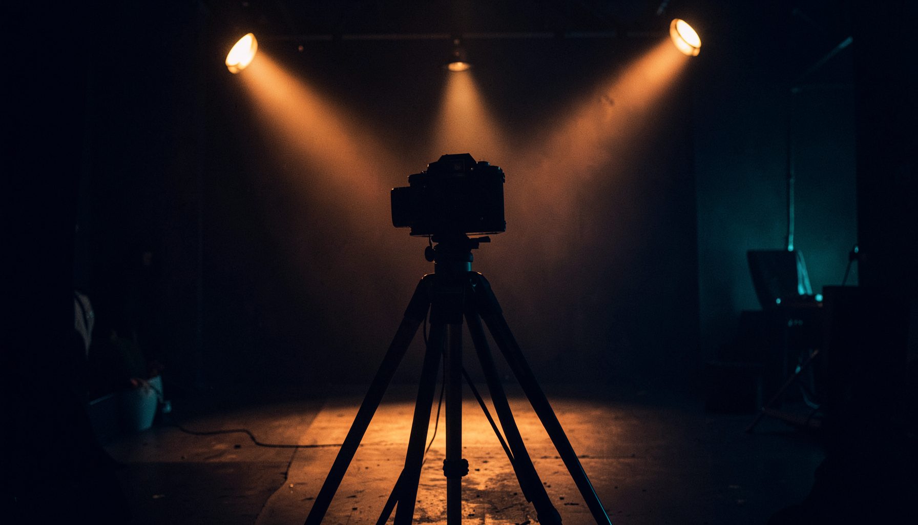 A dimly lit film set with a vintage camera on a tripod silhouetted against warm amber stage lights, deep navy shadows